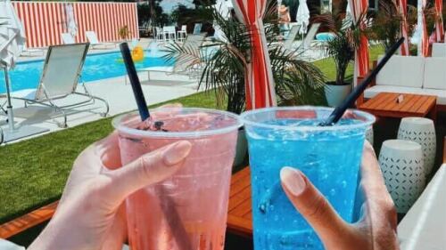 Two hands cheers drinks by a poolside. One of the drinks is a light pink and the other is an electric blue.