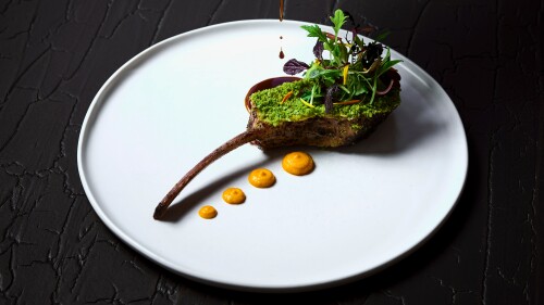 Sauce is poured over a decorated lamb chop on a white plate on a dinner table.