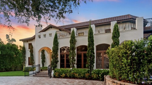 Mediterranean-style house with arched windows, tall cypress trees, and a sunset sky in the background.