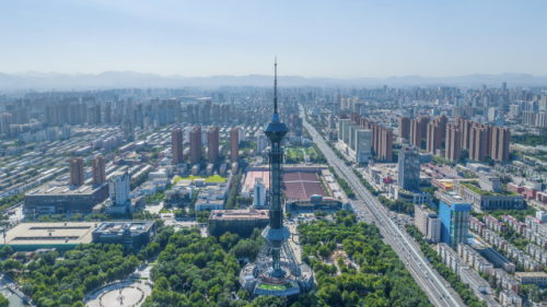 Aerial view of a cityscape with a prominent tall tower at the center surrounded by greenery. Buildings stretch into the distance under a clear blue sky.