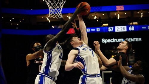 Duke and Louisville basketball players reach for a loose ball beneath a net in Spectrum Center.
