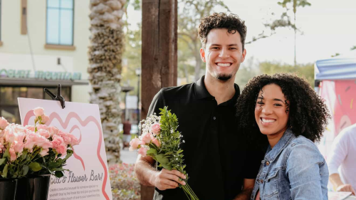 A smiling young man and woman stand together at an outdoor "Cupid's Flower Bar," holding a fresh bouquet of roses.