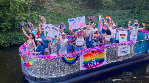 A barge full of people holding signs is traveling down a river. The boat's exterior is covered in silver sparkles, and a rainbow sign reads, "love is love."
