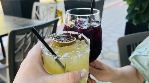 Two hands toast with a glass of margarita and a glass of sangria on an outdoor patio.