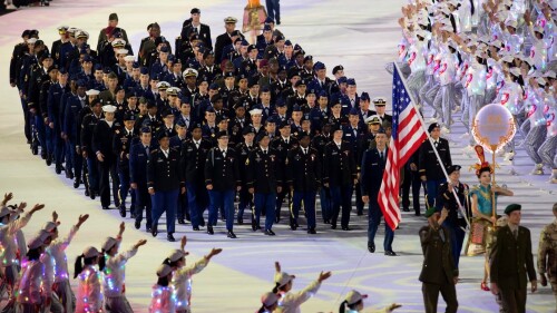A group of military members walking in formation in an Olympic-style venue, being cheered by other groups. 