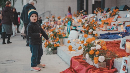 A young child wearing a colorful knit hat and dark jacket stands in front of a Día de los Muertos altar adorned with marigolds, candles, and framed photos. The altar is decorated with vibrant fabrics and marigold arrangements, creating a festive and respectful atmosphere as people move in the background.