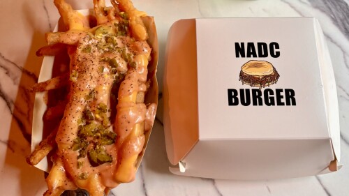 A tray of crinkle-cut fries covered in melted cheese, diced jalapeños, pepper, and a creamy orange sauce sits next to a white burger box labeled “NADC BURGER” with an illustration of a dripping cheeseburger on top. Both items are on a marble surface.