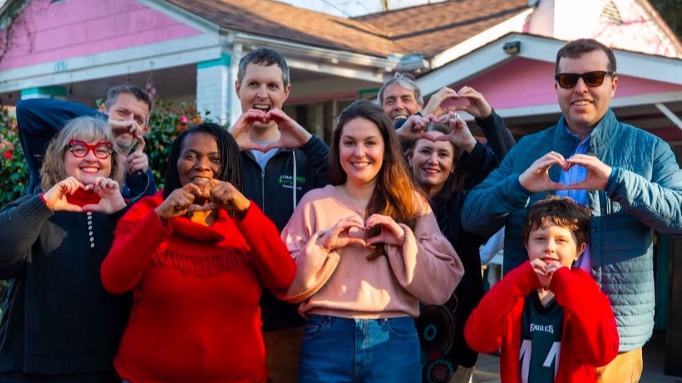 A diverse group of people stands smiling in front of a pink house. They all form heart shapes with their hands, conveying a warm, friendly atmosphere.