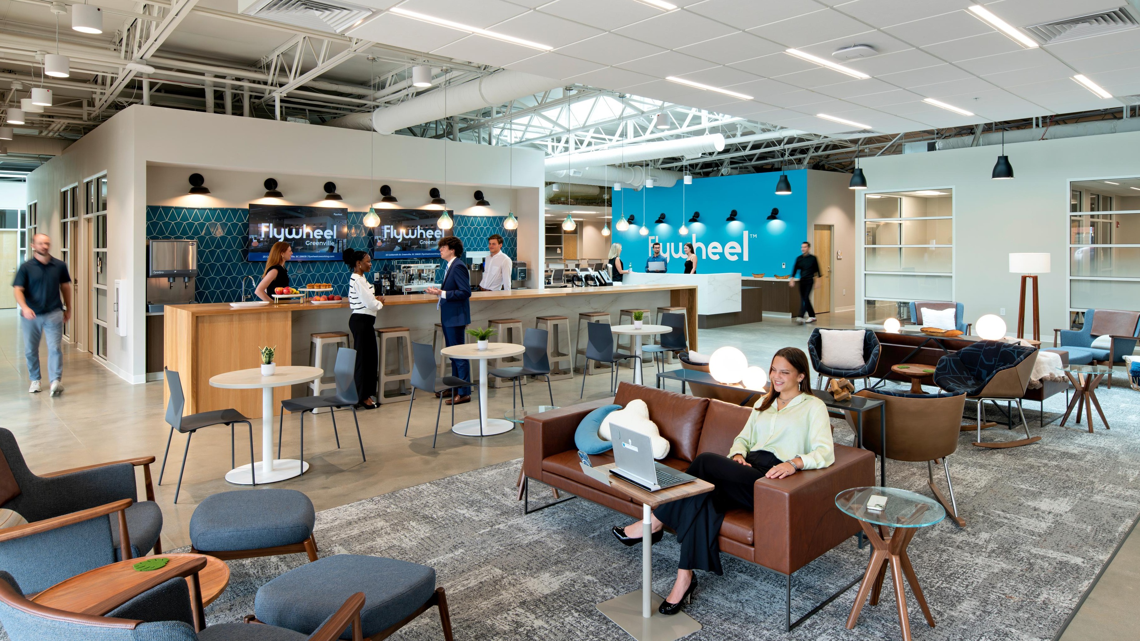 A coworking space with tables, chairs, a bar, someone working on a laptop, and a few people talking. 
