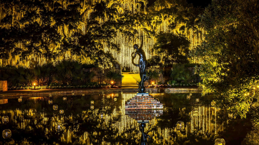 Golden lights drape from towering trees, casting a magical glow over a reflective pond. A graceful statue stands on a brick pedestal, mirrored in the water, surrounded by glowing lanterns.