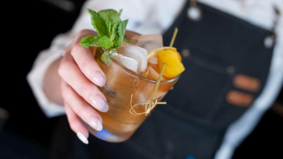 Bartender in a black apron holds an iced cocktail garnished with fresh mint and orange, conveying a refreshing, stylish bar atmosphere.