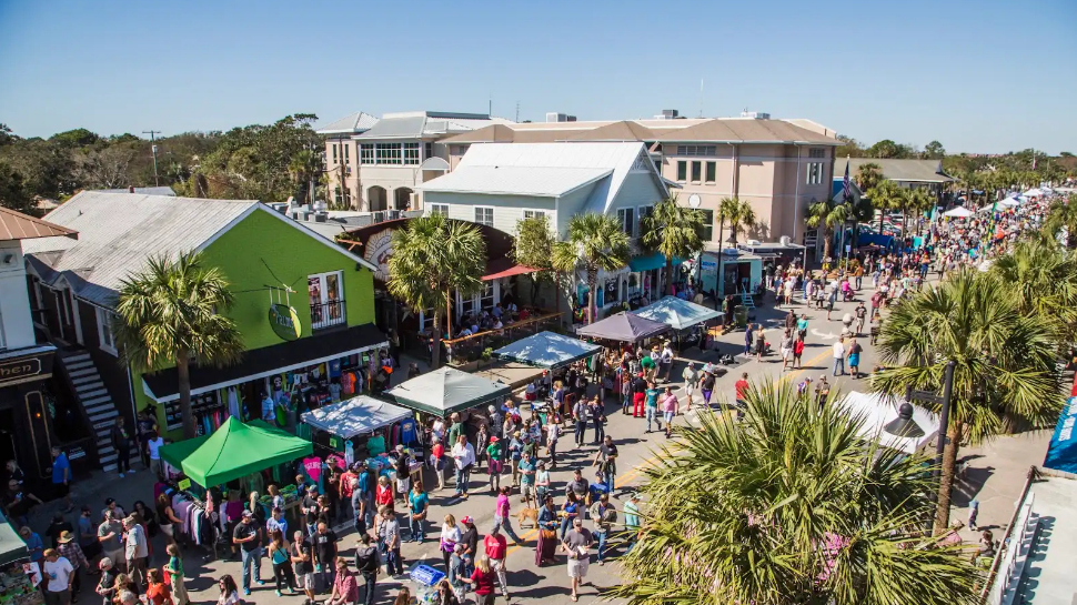 Aerial view of a lively street festival in a sunny coastal town, with colorful shops, palm trees, market stalls, and dense crowds.