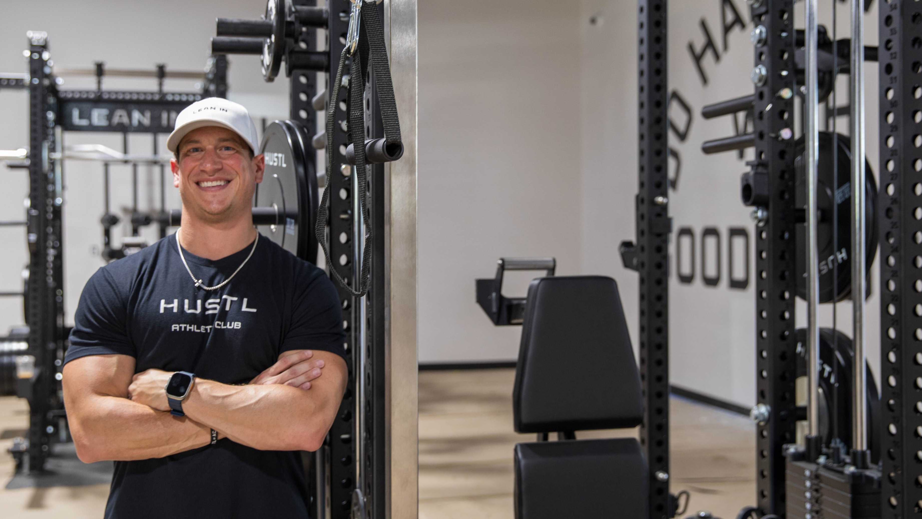 A person stands with arms crossed, smiling in a gym setting filled with weightlifting equipment and a bench.