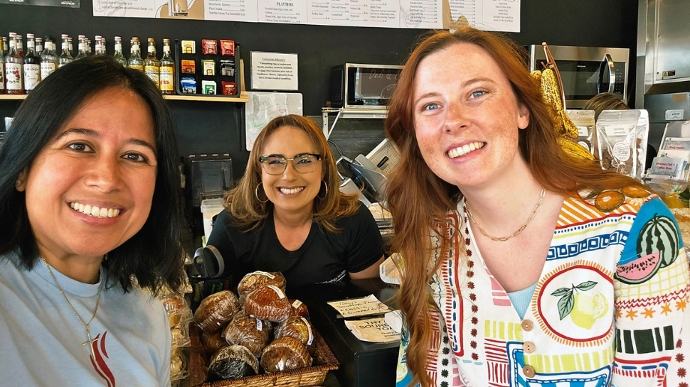 Three women smile behind a cafe counter stocked with muffins and cookies, with a large menu board displayed on the wall.