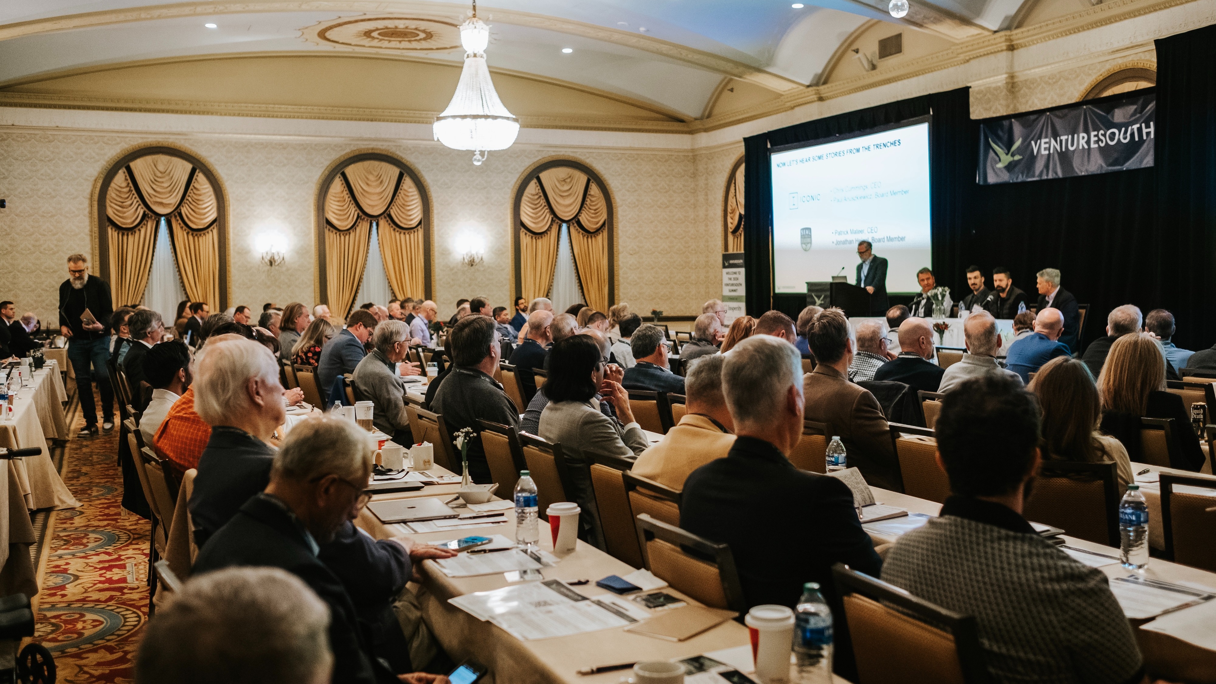 Conference room with people seated at long tables and a stage with panelists featuring a Venture South banner