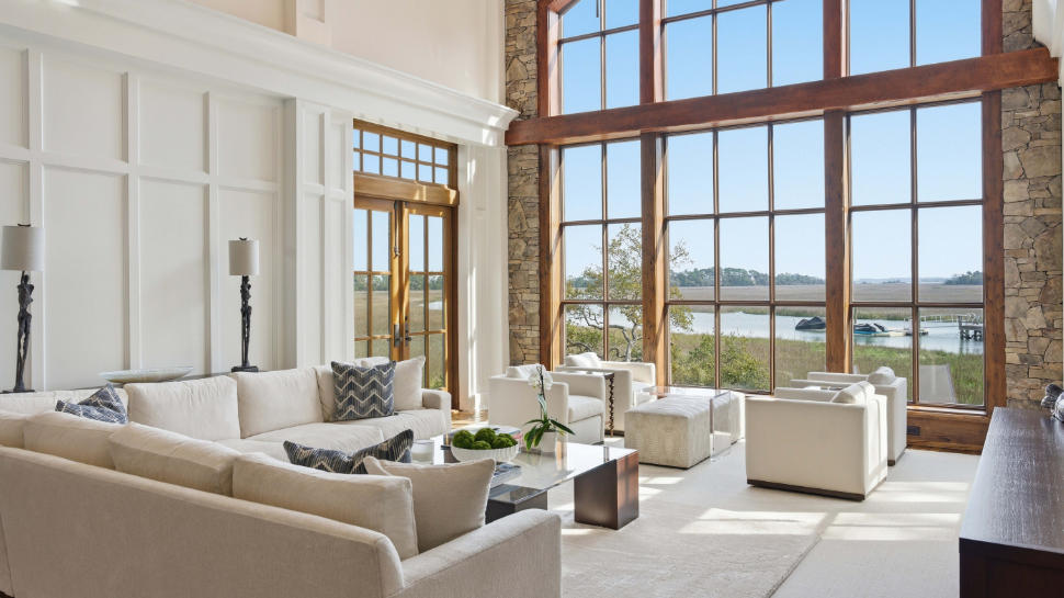 Bright modern-rustic living room with sofas, armchairs, stone walls, and floor-to-ceiling windows overlooking a tranquil lake and trees.