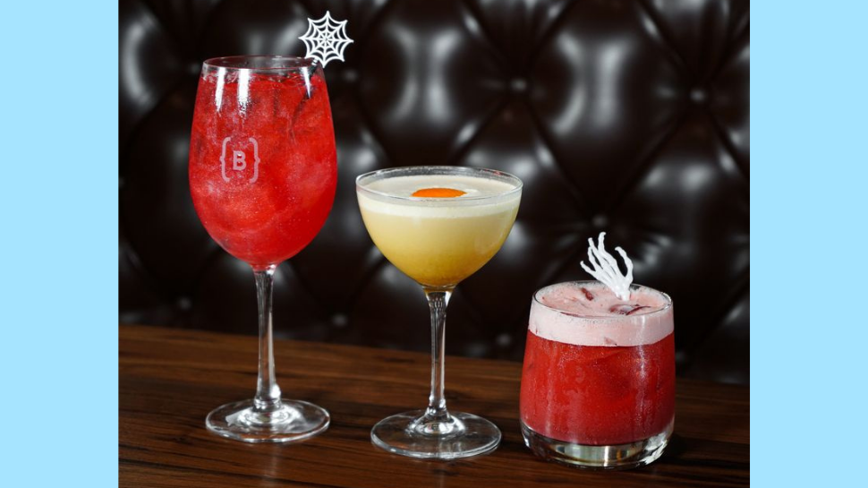 Three colorful cocktails—a red wine glass, yellow coupe, and red tumbler—sit on a wood table with spiderweb garnish, creating a festive mood.